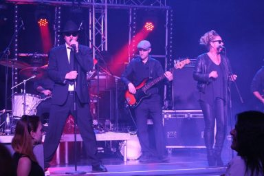 Eine Band tritt auf, mit Sängern in schwarzer Kleidung und Bandmitgliedern am Hintergrund.