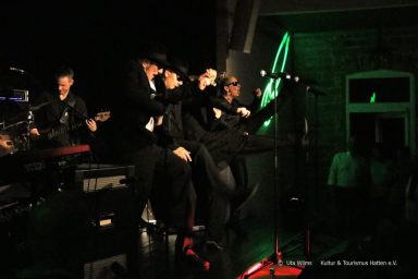 Bandmitglieder performen auf der Bühne mit Instrumenten und Beleuchtung im Hintergrund.